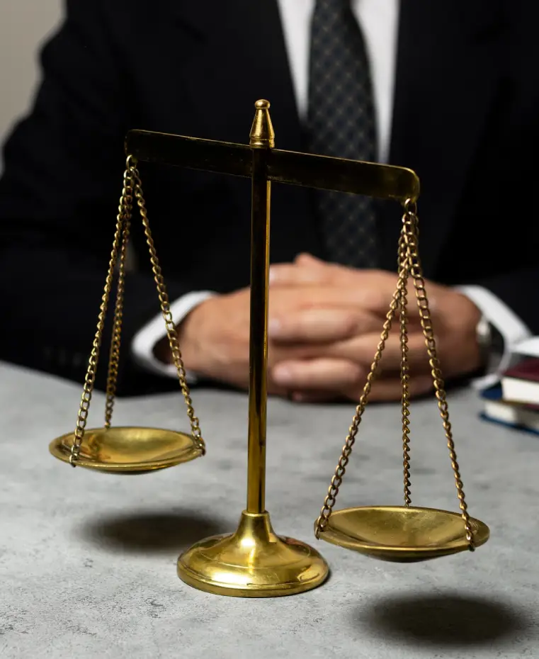 A golden scales of justice on a gray table, with a suited person’s hands clasped in the background, symbolizing legal representation